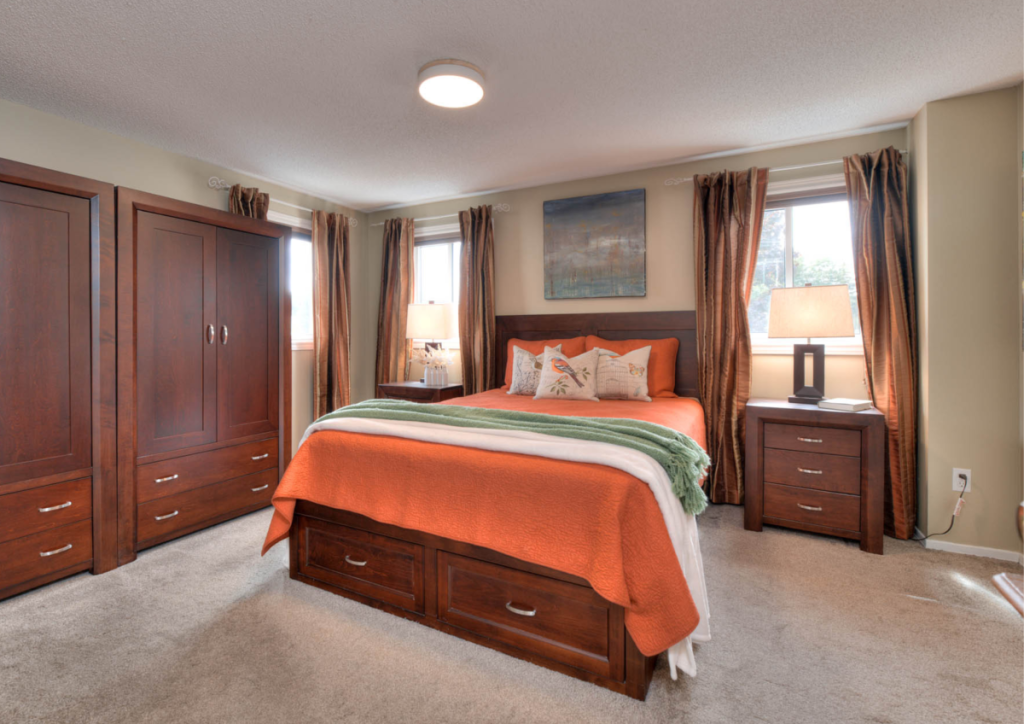 A staged bedroom with orange bedding, brown cabinetry, brown curtains, and warm-toned accents by Helen’s Team.